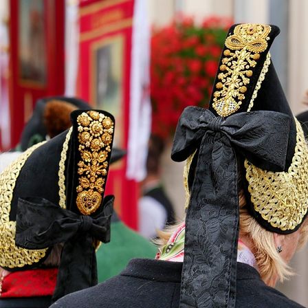 Zwei Personen in traditioneller Kleidung stehen mit Blick nach hinten, mit aufwendigen gold- und schwarzfarbenen Hüten, die mit Schleifen verziert sind, möglicherweise bei einer Parade. Der unscharfe Hintergrund zeigt eine rote Wand mit einem Bilderrahmen und roten Blumen.