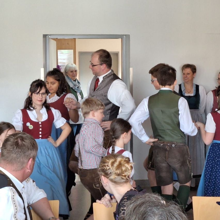 Eine Gruppe von Menschen in traditioneller Kleidung, die in einem Raum mit weißen Wänden und einer Holztür stehen und sich unterhalten. Erwachsene und Kinder sind anwesend.