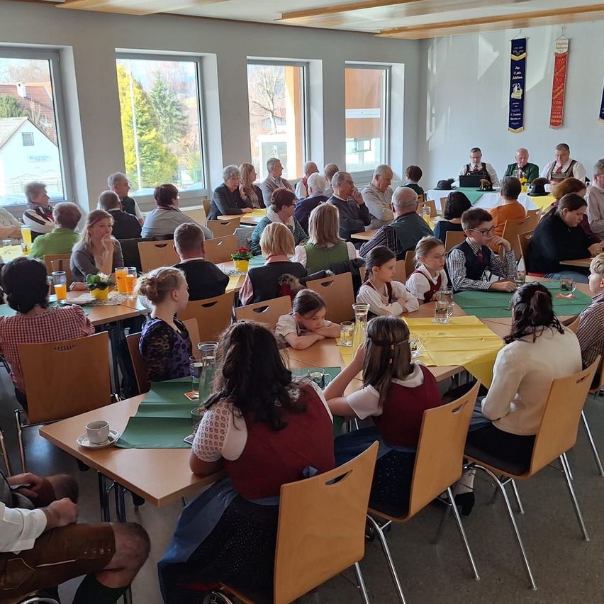 Eine große Gruppe von Menschen sitzt in einem Speisesaal, einige unterhalten sich, während andere etwas betrachten. Der Raum hat mehrere Tische mit gelben Tischdecken und Stühlen. Es gibt Menschen, die Brillen tragen, und einige tragen Hüte. An der Wand hängen Banner und Fenster.