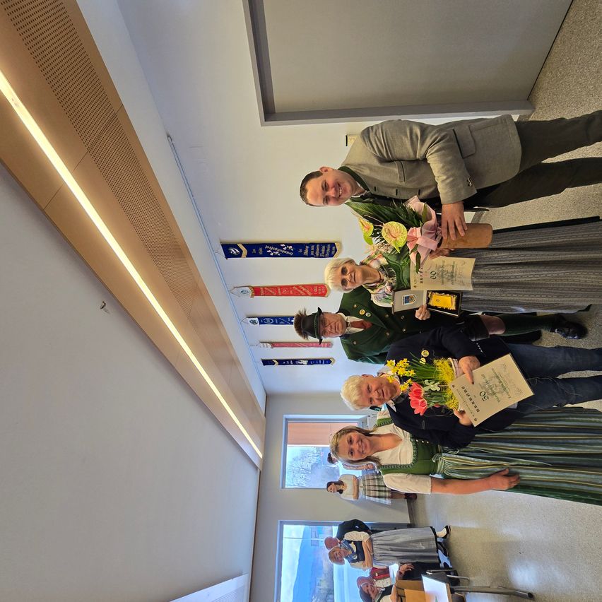 Eine Gruppe von Menschen in traditioneller Kleidung posiert für ein Foto in einem Flur mit Bannern und einem Fenster im Hintergrund.