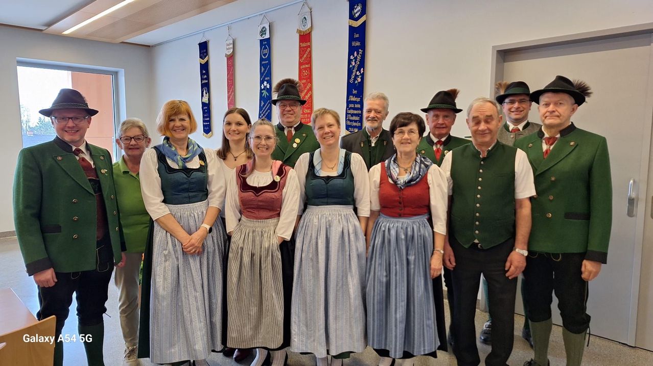 Eine Gruppe von Menschen in traditioneller Kleidung steht in einem Raum mit weißen Wänden und Bannern hinter ihnen. Sie lächeln und posieren für ein Foto.
