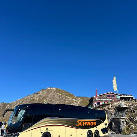 Ein großer, gelber Bus mit orangefarbenem Text ist auf einer Bergstraße geparkt, mit einem Gebäude und Fahnen dahinter.