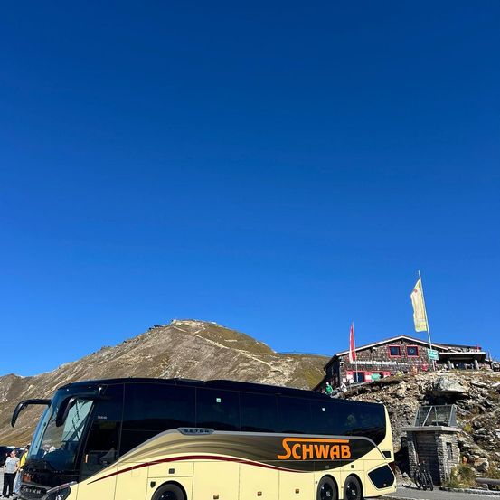 Ein großer, gelber Bus mit orangefarbenem Text ist auf einer Bergstraße geparkt, mit einem Gebäude und Fahnen dahinter.