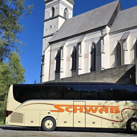Ein Bus mit dem Wort Schwab darauf parkt vor einer Kirche mit aufwendigen Verzierungen an ihren Wänden.