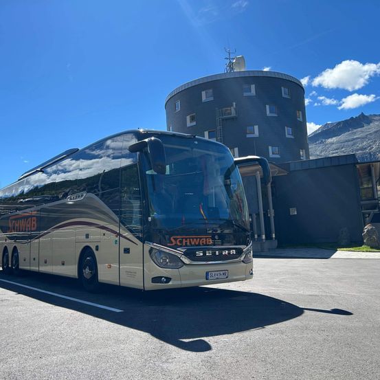 Ein großer weißer Bus mit der Aufschrift SCHWAB SETRA ist vor einem modernen Gebäude geparkt. Die Türen des Busses sind geöffnet und der Schatten des Busses fällt auf die Straße. Ein Berg ist im Hintergrund sichtbar.
