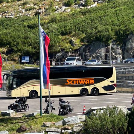 Ein Bus mit SCHWAB auf der Seite ist auf einer Straße geparkt, in der Nähe zweier Motorräder. Außerdem sind Autos auf einer Brücke geparkt und eine Fahne steht auf einem Pfosten.