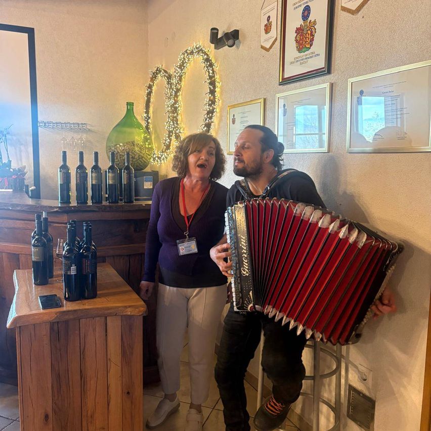 A woman laughs while a man plays an accordion in a room filled with wine bottles and framed certificates on the wall.