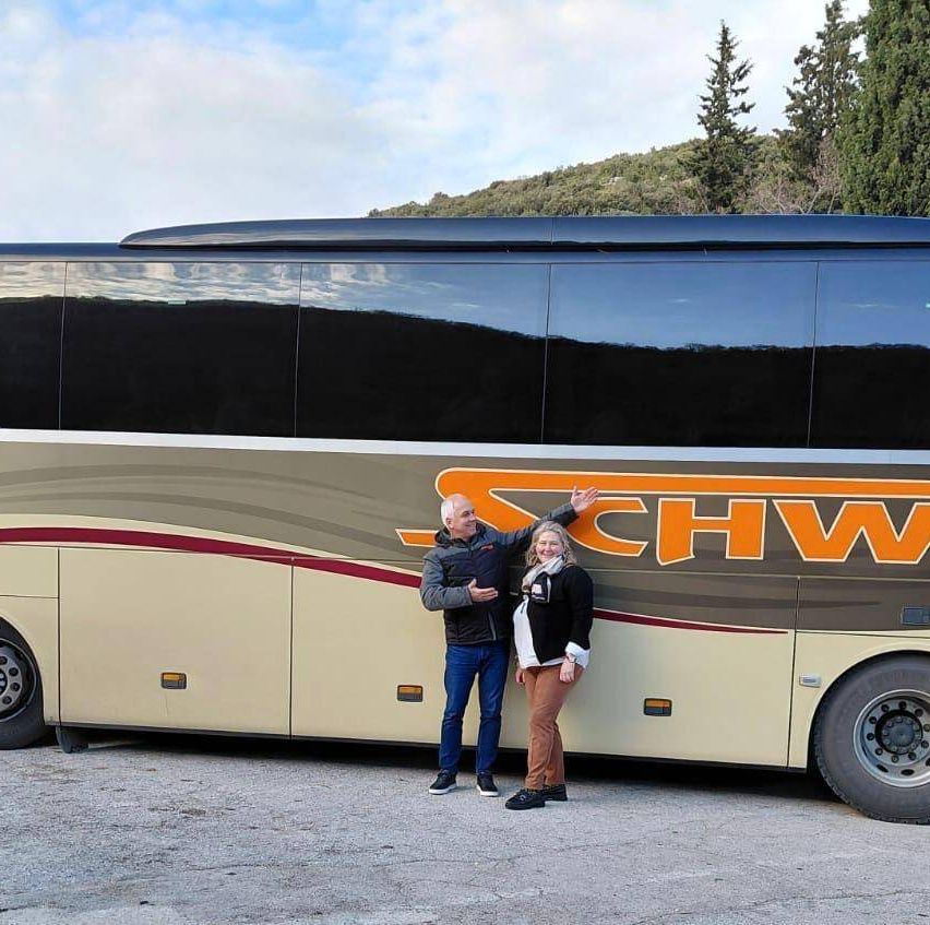 Two people, a man and a woman, are standing in front of a large bus with SCHW on the side, against a backdrop of trees and mountains.