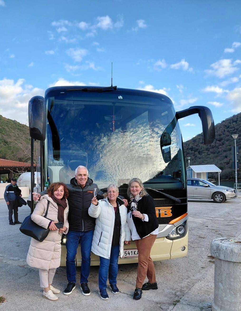 Three people, two women and a man, stand in front of a bus, holding wine glasses and smiling. Behind them, a mountain and a parked car are visible.