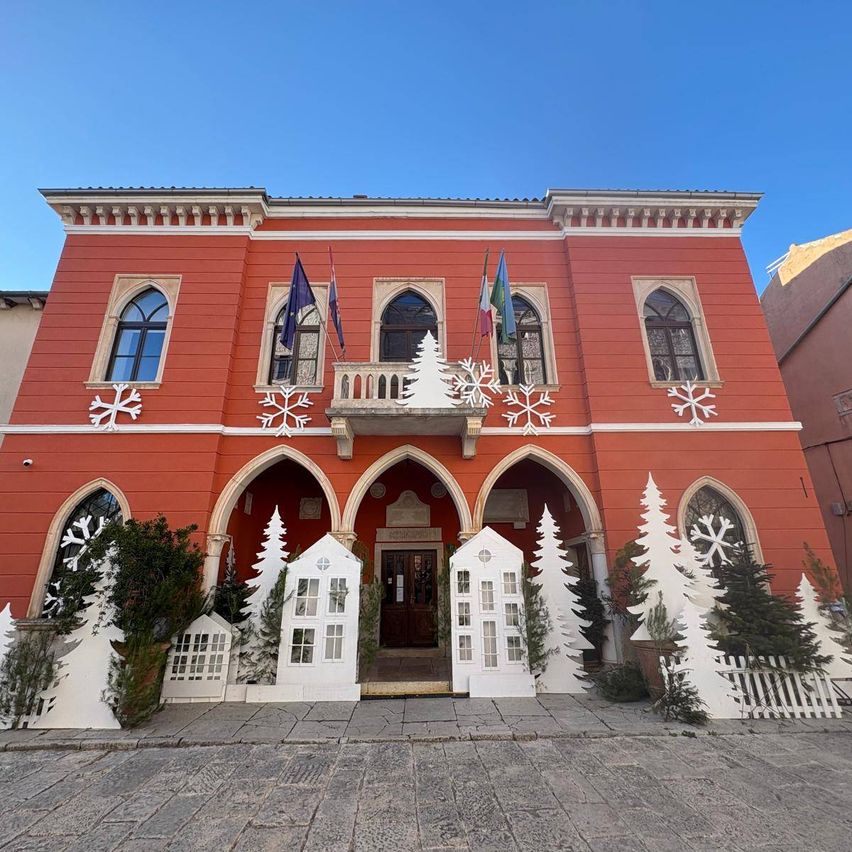 A red building with multiple arched windows and flags on the balcony is decorated with Christmas trees and snowflakes.