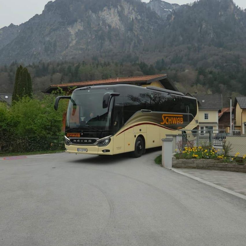 A yellow bus labeled Schwab is parked on the road with mountains and houses in the background.