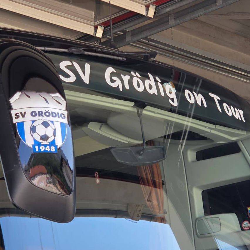 Nahaufnahme eines Busseitenspiegels mit einem blau-weißen Fußballteam-Logo und dem Text 'SV Grodig auf Tour'. Der Spiegel reflektiert ein Gebäude und den Himmel.