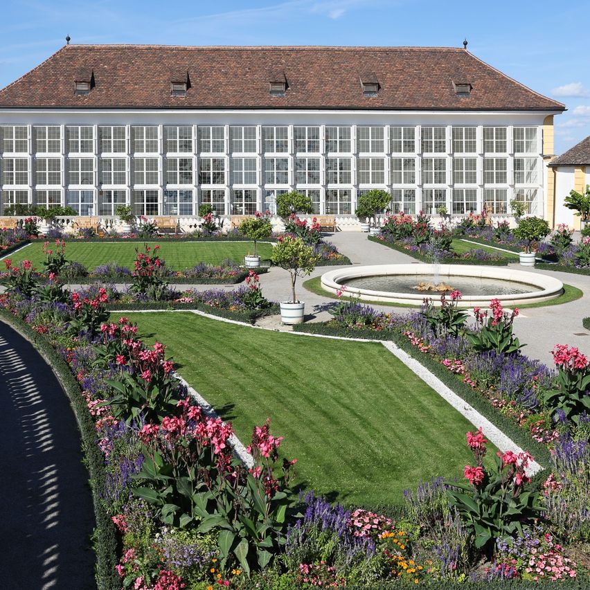 Ein großes Gebäude mit vielen Fenstern, umgeben von einem gut gepflegten Garten mit verschiedenen Blumen und einer Fontäne in der Mitte.