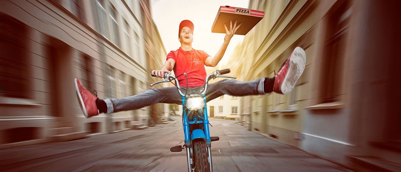Ein Pizzabote fährt mit einem blauen Motorrad, hält eine Pizzaschachtel und hat eine verschwommene Stadtstraße im Hintergrund.