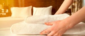 Eine Person faltet weiße Handtücher auf einem Bett mit Kissen im Hintergrund. Der Raum ist gut beleuchtet, wahrscheinlich durch ein Fenster.