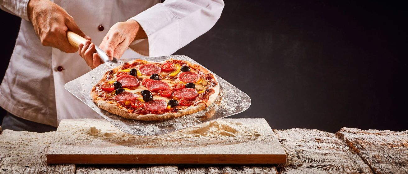 Ein Koch, der einen Pizzaschaufel hält, ist dabei, eine Pizza von einem Backblech zu nehmen. Die Pizza hat Beläge wie Peperoni, schwarze Oliven und Tomaten.