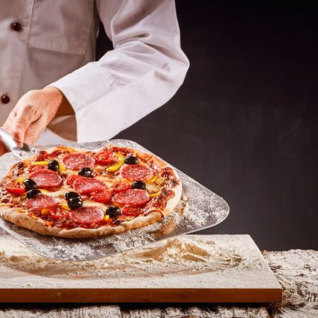 Ein Koch in weißer Uniform bereitet eine Pizza mit Peperoni und schwarzen Oliven auf einem bemehlten Metalltablett zu.