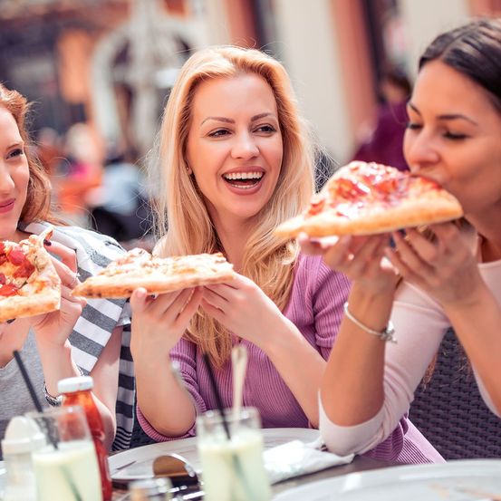 Drei Frauen genießen im Freien Pizzastücke und lachen und lächeln. Im Hintergrund ist eine verschwommene Gebäudeansicht und andere Menschen zu sehen.