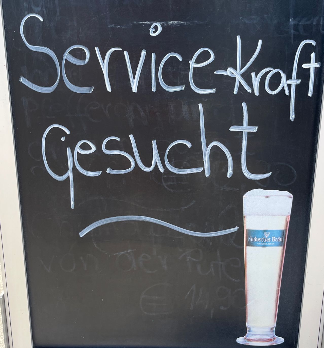 Ein Tafel mit der Aufschrift 'Servicekraft gesucht' (Servicekraft gesucht) ist zu sehen, mit einem Glas Bier mit einem blauen Etikett in der Ecke.