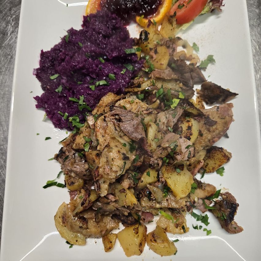 Ein weißer Teller mit einem Fleischgericht und lila Kartoffelpüree, garniert mit Kräutern und einer Tomatenscheibe.