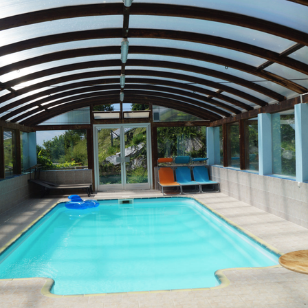 Ein Hallenbad mit durchsichtigem Dach, blauem Wasser und Liegestühlen. Der Pool ist von Glaswänden umgeben, und es gibt einen kleinen Tisch und einen Liegestuhl.