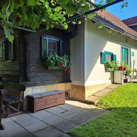 Ein kleines Haus mit braunem Dach und grünen Fensterläden. Blumentöpfe schmücken die Fenster. Eine Holzbank und eine Aufbewahrungsbox sind auf dem Patio.