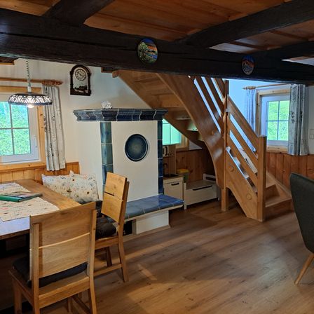 Ein gemütlicher Essbereich mit Holzmöbeln, einem Tisch, Stühlen, einer Bank und einem Kamin. Der Raum hat Holzböden, Wände und eine Decke mit sichtbaren Balken. Es gibt Fenster mit Vorhängen und eine Lampe, die von der Decke hängt.