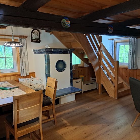 Ein gemütlicher Essbereich mit Holzmöbeln, einem Tisch, Stühlen, einer Bank und einem Kamin. Der Raum hat Holzböden, Wände und eine Decke mit sichtbaren Balken. Es gibt Fenster mit Vorhängen und eine Lampe, die von der Decke hängt.