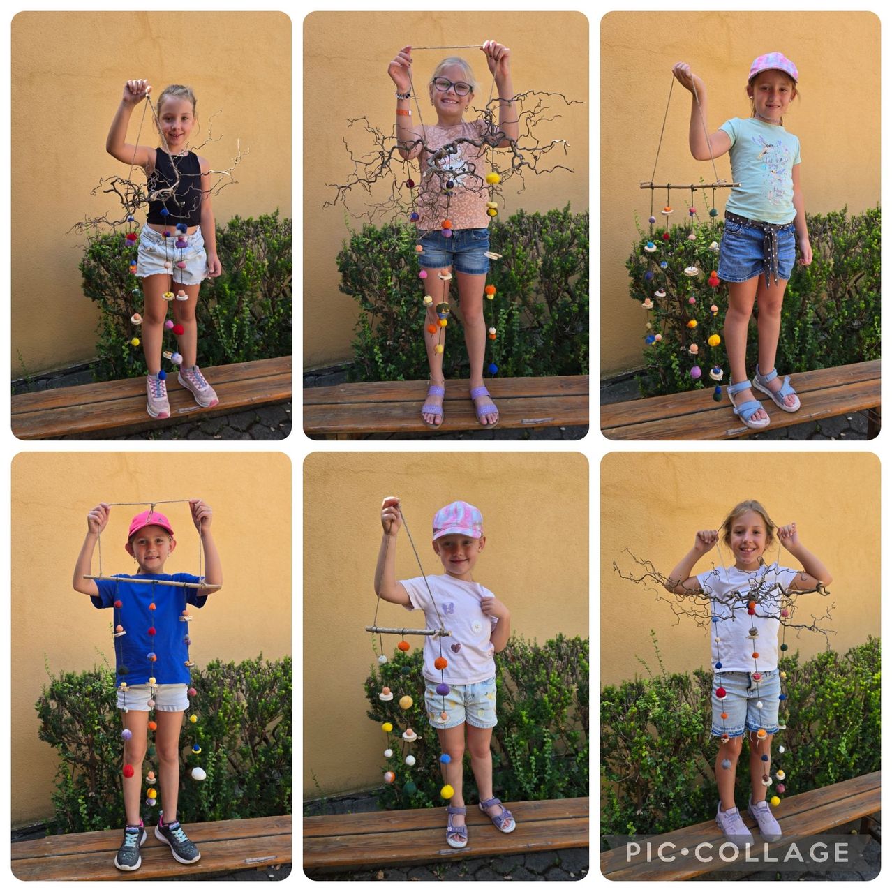Six children, each holding a colorful, handmade craft made of twigs and pom-poms, stand on wooden benches against a yellow wall.