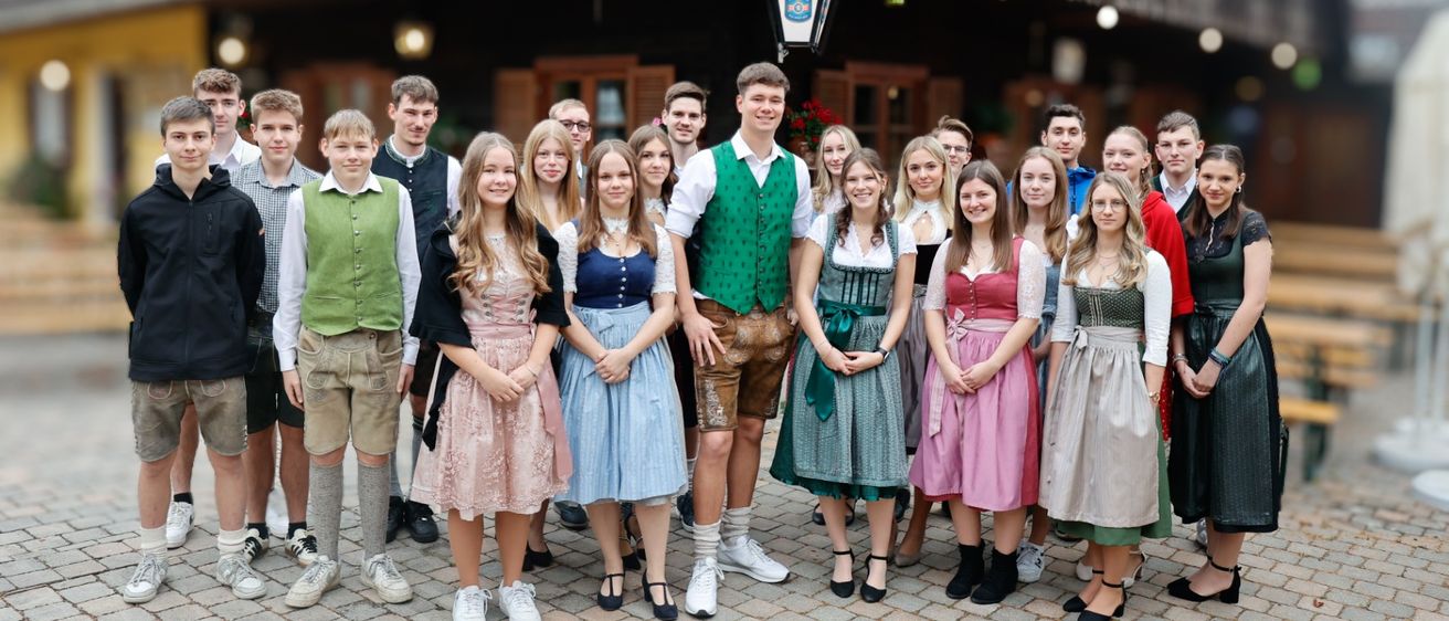 Eine Gruppe junger Menschen in traditioneller deutscher Kleidung, einschließlich Lederhosen und Dirndln, posiert für ein Foto vor einem rustikalen Gebäude.