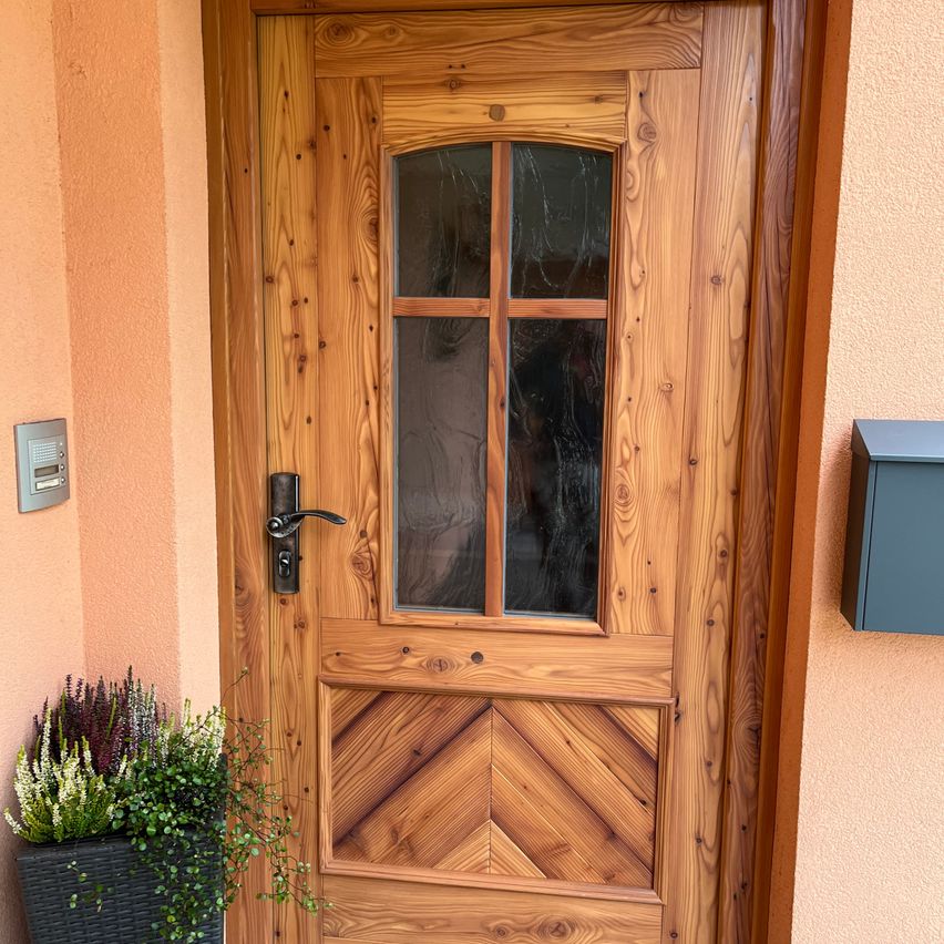 Eine Holztür mit einem Glasfenster in der Mitte und einem Metallgriff ist an einer beigefarbenen Wand befestigt. Links steht eine Pflanze.