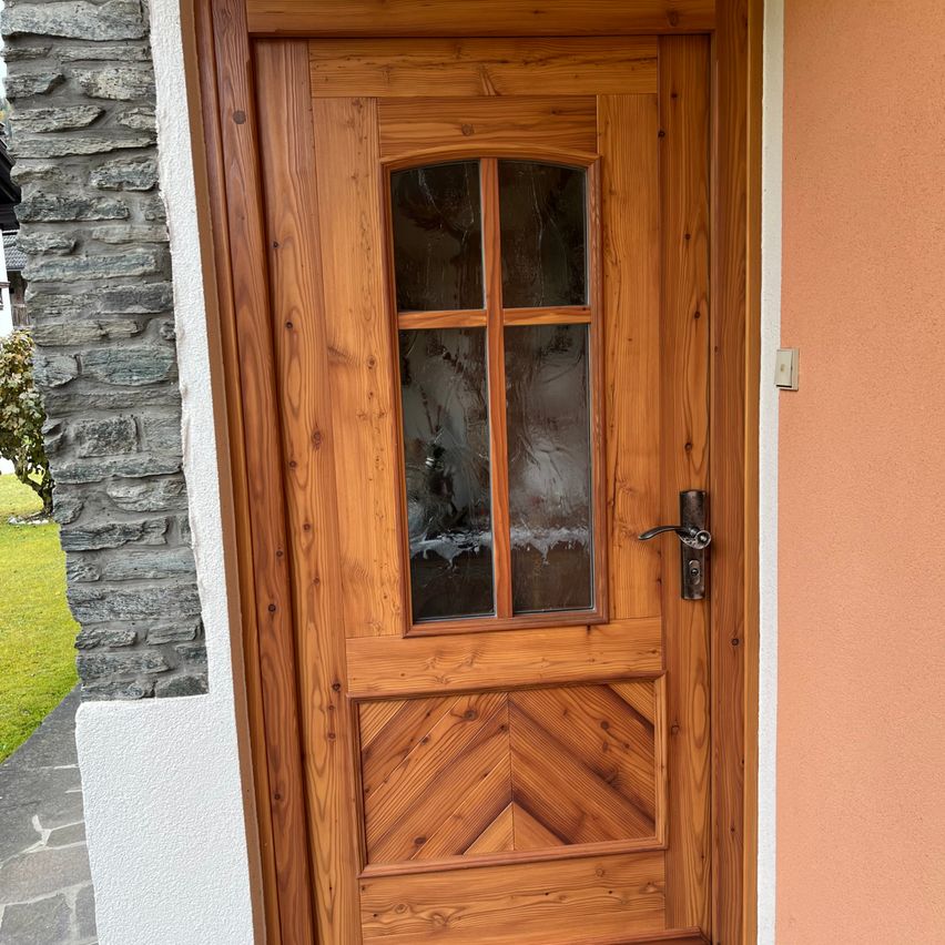 Eine Holztür mit Glasscheiben ist an einer beigen Wand montiert. Die Tür hat einen Holzgriff und ein gemustertes Design im unteren Teil.