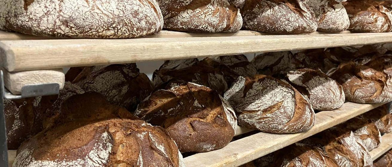 Mehrere Regale, gestapelt mit Artisan-Brot, präsentieren eine Vielzahl von Laibe mit strukturierter Kruste, einige mit sichtbaren Schnitten, gelagert in einer Bäckerei.