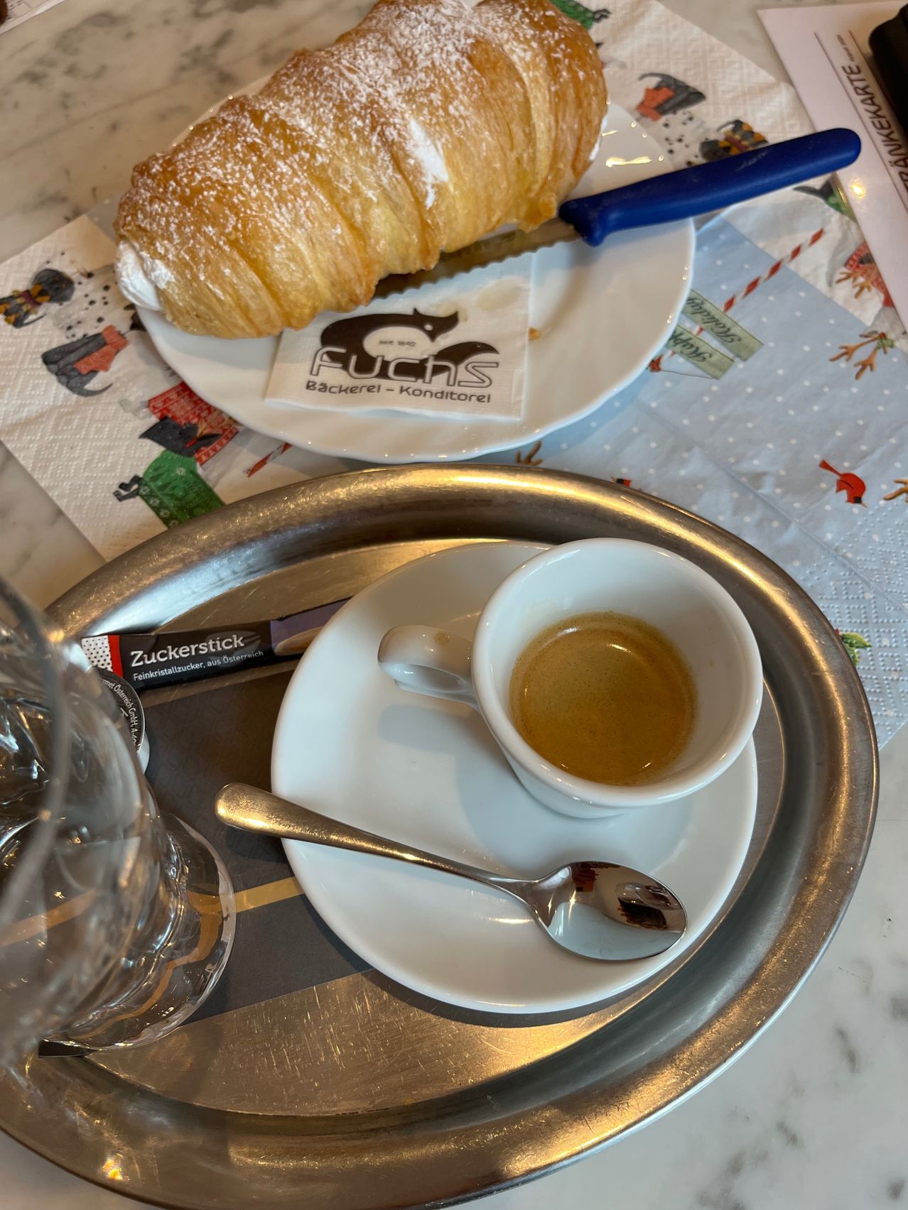 A croissant sits on a white plate with a knife, while a cup of espresso rests on a saucer with a spoon, on a silver tray with a glass of water.