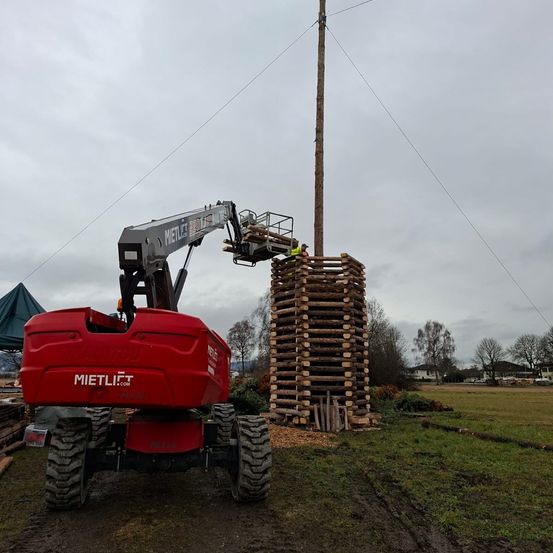 Ein roter Kran hebt Holzstapel zu einem hohen Holzpfahl. Eine Person ist auf dem Kran. Der Himmel ist bewölkt.