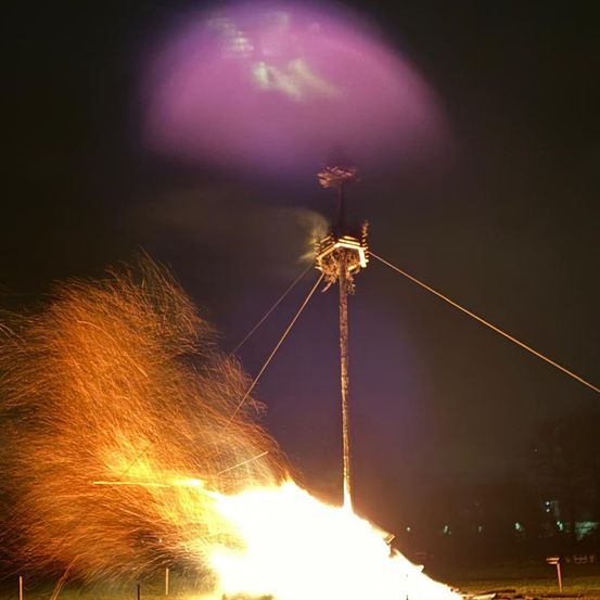 Ein Feuerwerk mit einer hellen gelben Flamme und Funken, die sich in der Luft ausbreiten, gegen einen dunklen Nachthimmel mit einem lila Schimmer.