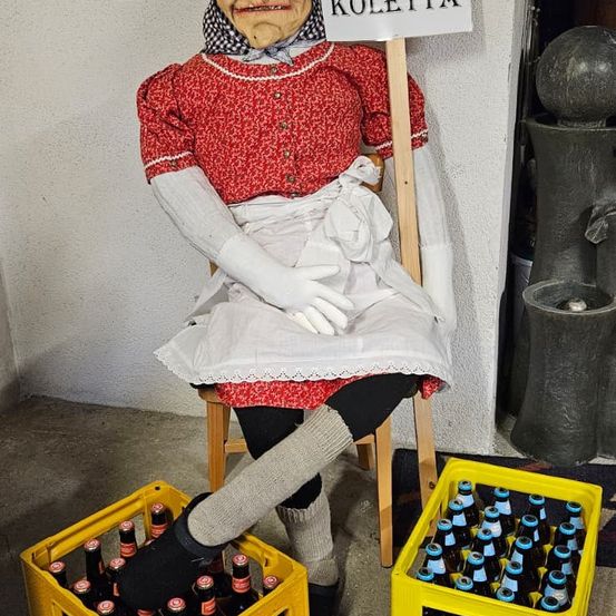 Eine Puppe, gekleidet wie eine alte Frau namens Koletta, sitzt in einem Stuhl mit zwei gelben Kisten voller Flaschen davor. Das Schild auf dem Stuhl lautet Koletta. Hinter dem Stuhl steht eine graue Zement-Skulptur.