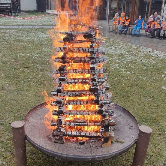 Ein hoher Stapel brennender Holzscheite in einem Metall-Feuerkorb, umgeben von Kindern in orangefarbenen Westen.