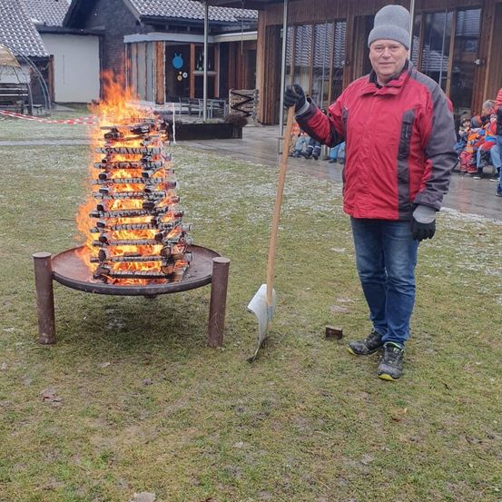 Ein Mann steht neben einem brennenden Lagerfeuer und hält eine Schaufel. Das Feuer ist von einem Haufen Holz umgeben.