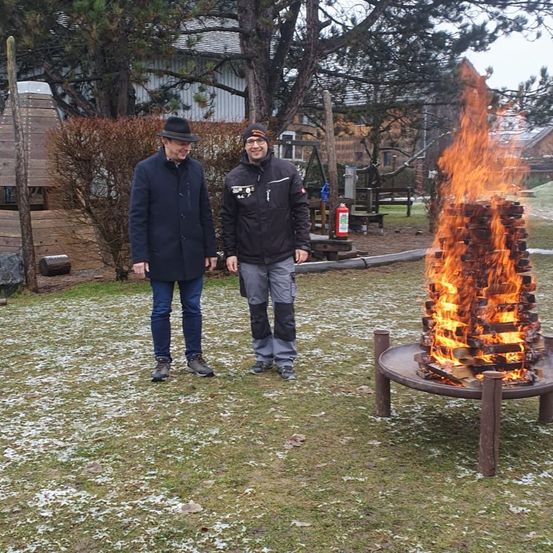 Zwei Männer stehen in einem Park, einer trägt einen Hut und der andere eine Mütze. Sie stehen neben einem brennenden Feuerkorb.