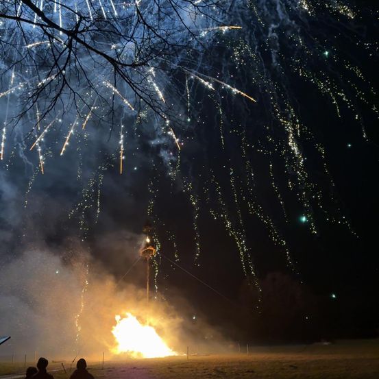 Ein großes Lagerfeuer mit explodierenden Feuerwerken darüber, zwei Personen beobachten von unten. Bäume ohne Blätter umgeben die Szene.