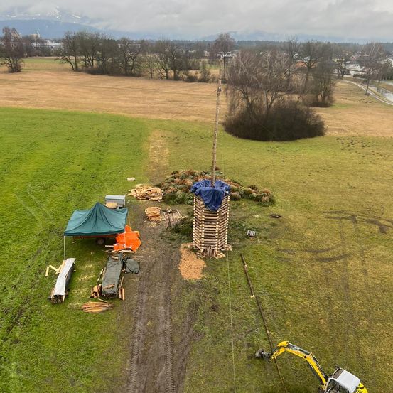 Luftaufnahme einer ländlichen Landschaft mit einem Feld, einem hohen Pfahl, einem Holzstapel, einem Zelt und einem gelben Bagger.