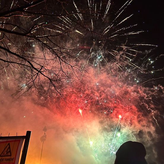 Ein farbenfroher Feuerwerksdisplay erleuchtet den Nachthimmel, wobei sich der Rauch in mehreren Farben ausbreitet. Unten ist die Silhouette einer Person mit Hut zu sehen. Ein Schild im Vordergrund warnt vor Feuerwerk.