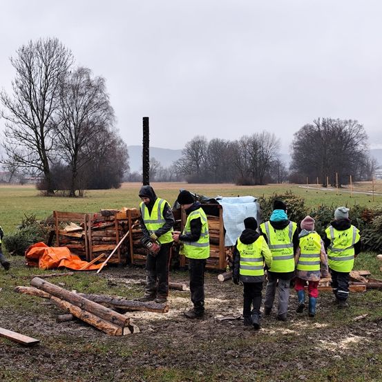 Mehrere Personen in Warnwesten stehen auf einem Grasfeld, mit einem Stapel Holz und einer großen Plane in der Nähe. Im Hintergrund säumen Bäume die Landschaft, und Berge sind am Horizont sichtbar.