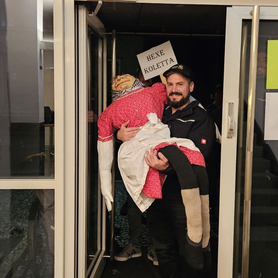 Ein Mann hält eine Puppe, lächelt und trägt sie durch eine Tür. Die Puppe hat ein Schild mit der Aufschrift 'Hexe Koletta' auf der Brust.