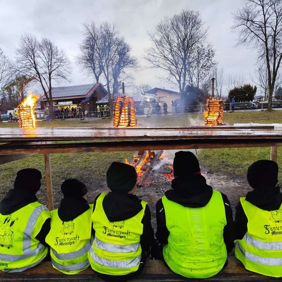 Vier Personen in Warnwesten sitzen in der Nähe eines Lagerfeuers, mit zusätzlichen brennenden Feuern auf einem Tisch vor ihnen und einem bewölkten Himmel im Hintergrund.