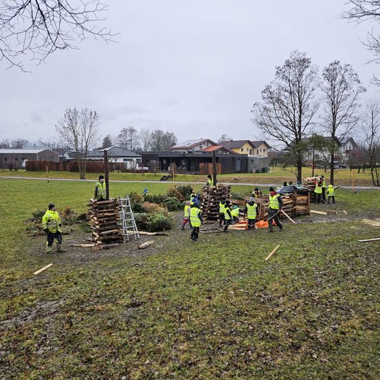 Eine Gruppe von Menschen in gelben Westen arbeitet auf einem Rasen, umgeben von mehreren Häusern. Einige stapeln Holz, andere schneiden es.