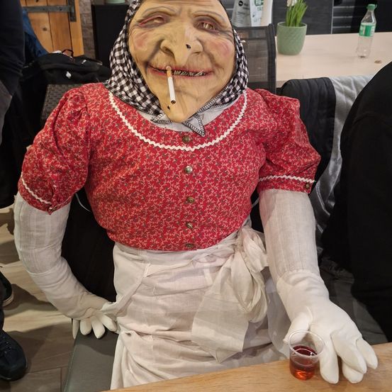 Eine Person in einer Maske mit einer Pfeife im Mund, die ein rotes Kleid und eine weiße Schürze trägt, sitzt an einem Tisch mit einem Getränk.