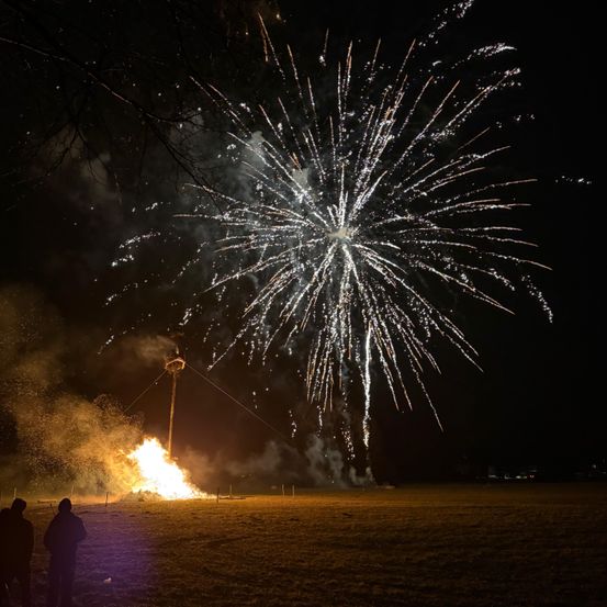 Ein Feuerwerk bei Nacht, bei dem zwei Personen aus der Ferne zusehen und ein Feuer im Vordergrund brennt.