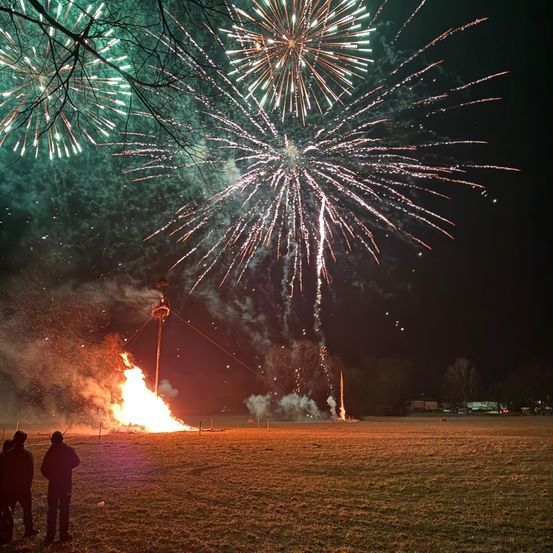 Feuerwerke explodieren in der Nacht mit einem großen Lagerfeuer in der Mitte. Zwei Personen stehen vor dem Feuer.